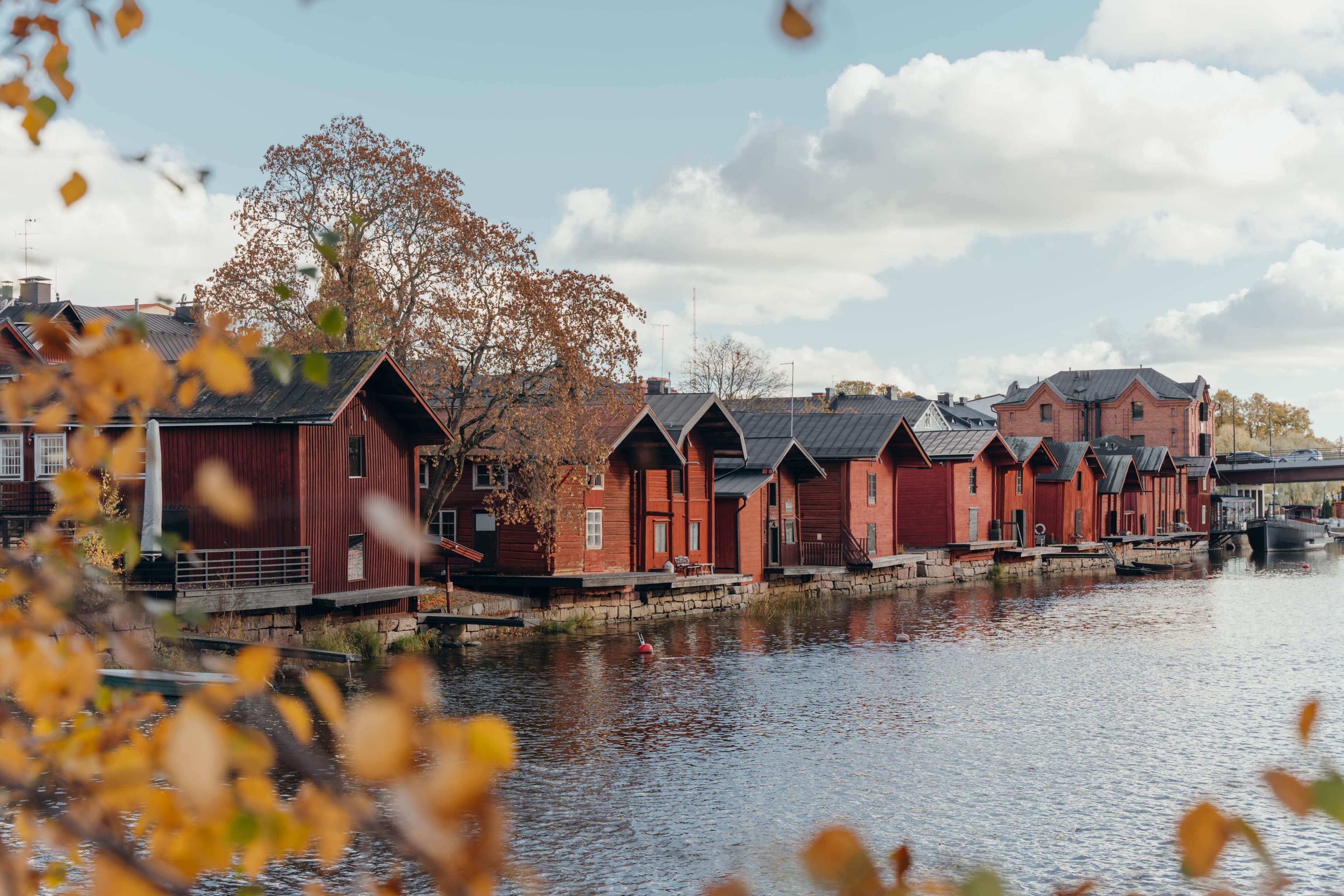 Row of red wooden houses by a tranquil river under a cloudy sky, framed by autumnal foliage.